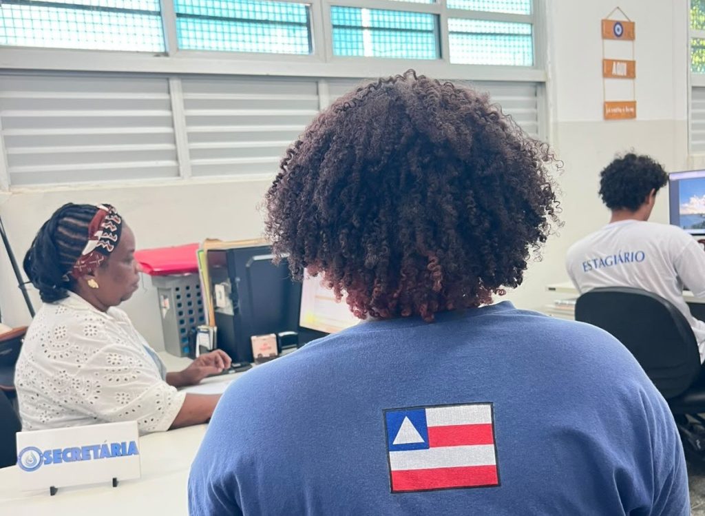 Pessoa de costas, com camiseta azul e bandeira da Bahia, diante de um balcão identificado como secretaria; outras pessoas trabalham em computadores.