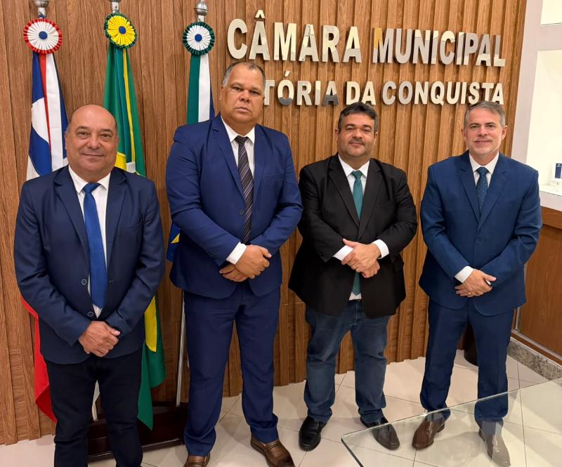 Quatro homens de terno em frente ao painel da Câmara Municipal de Vitória da Conquista e a bandeiras
