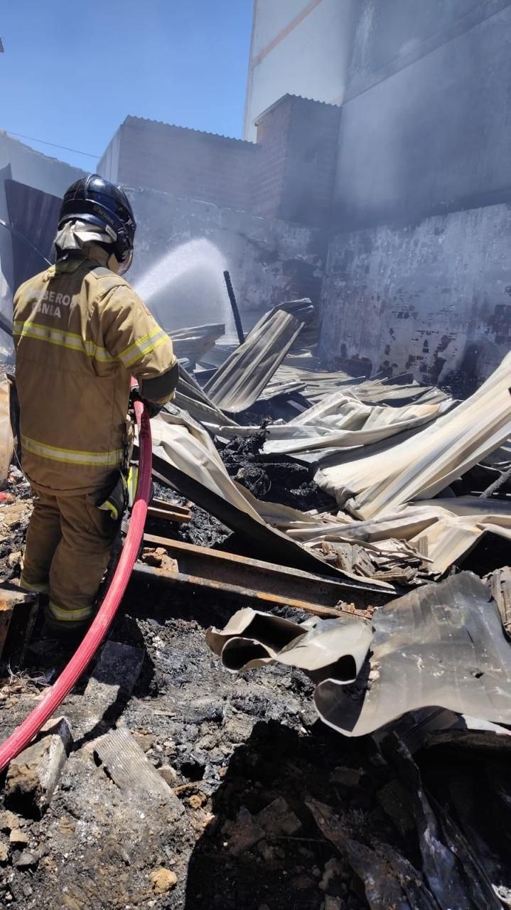Bombeiro com equipamentos de proteção usa mangueira em área com destroços, telhas retorcidas e fumaça