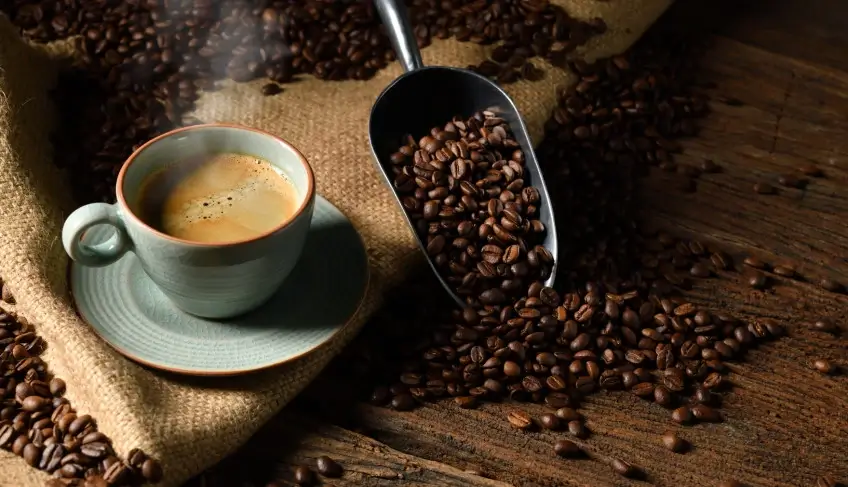 Xícara de café ao lado de grãos torrados e uma pá sobre uma mesa. Imagem ilustrativa.