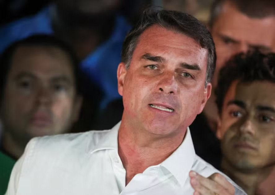 Flávio Bolsonaro de camisa branca, com outras pessoas ao fundo.