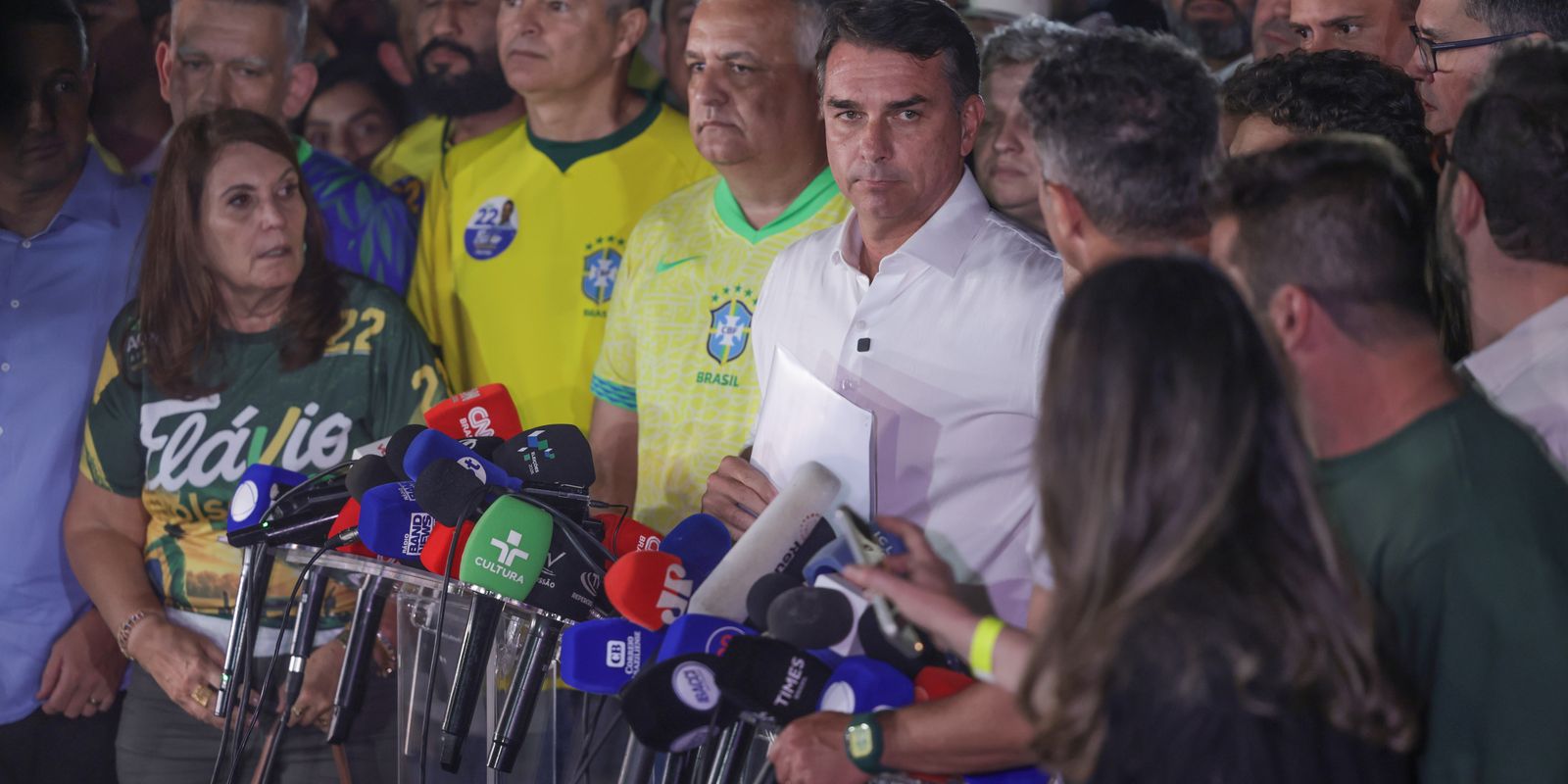 Flávio Bolsonaro diante de microfones e apoiadores em entrevista à imprensa.