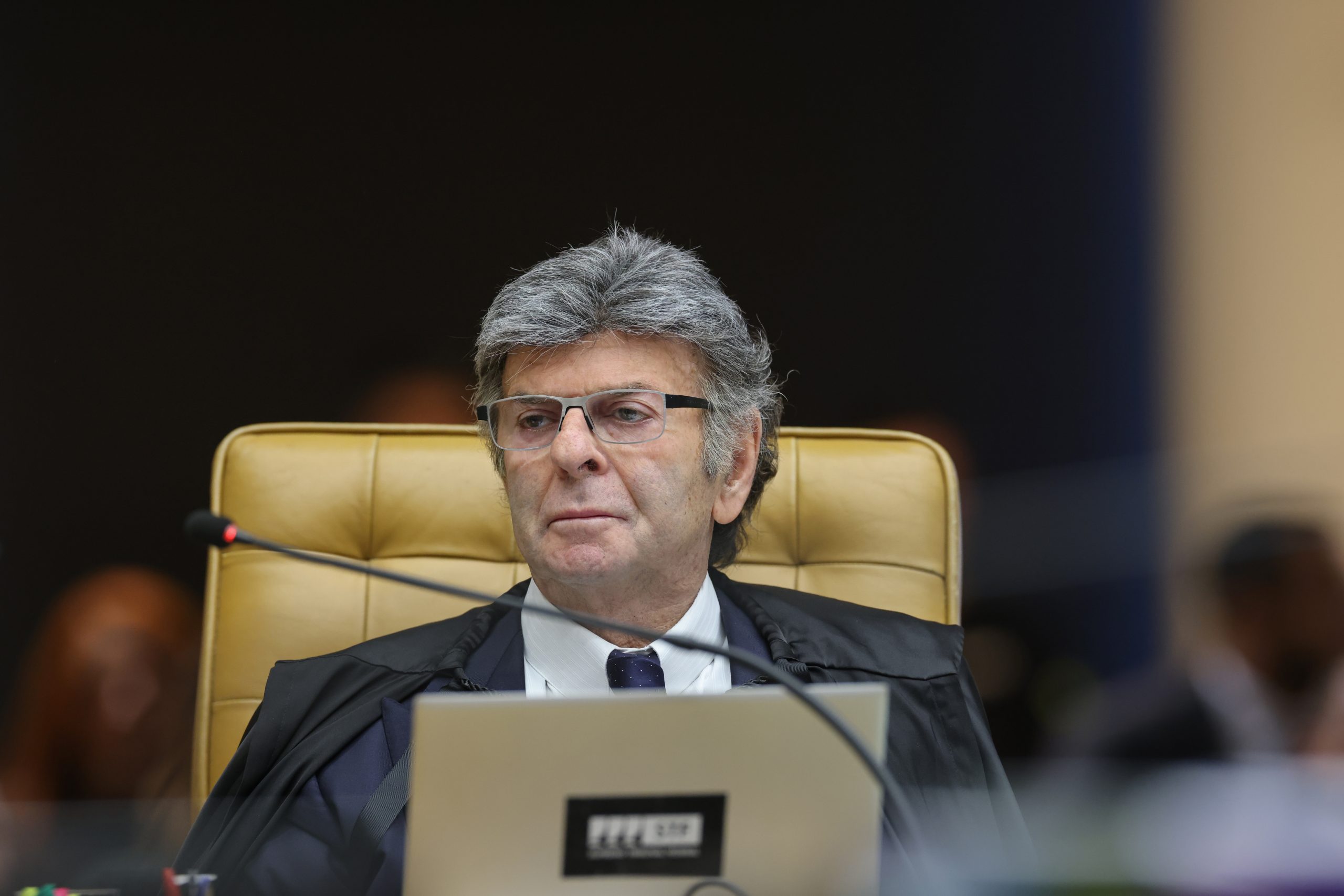 Luiz Fux sentado em cadeira do plenário do STF, com óculos e toga, diante de um computador