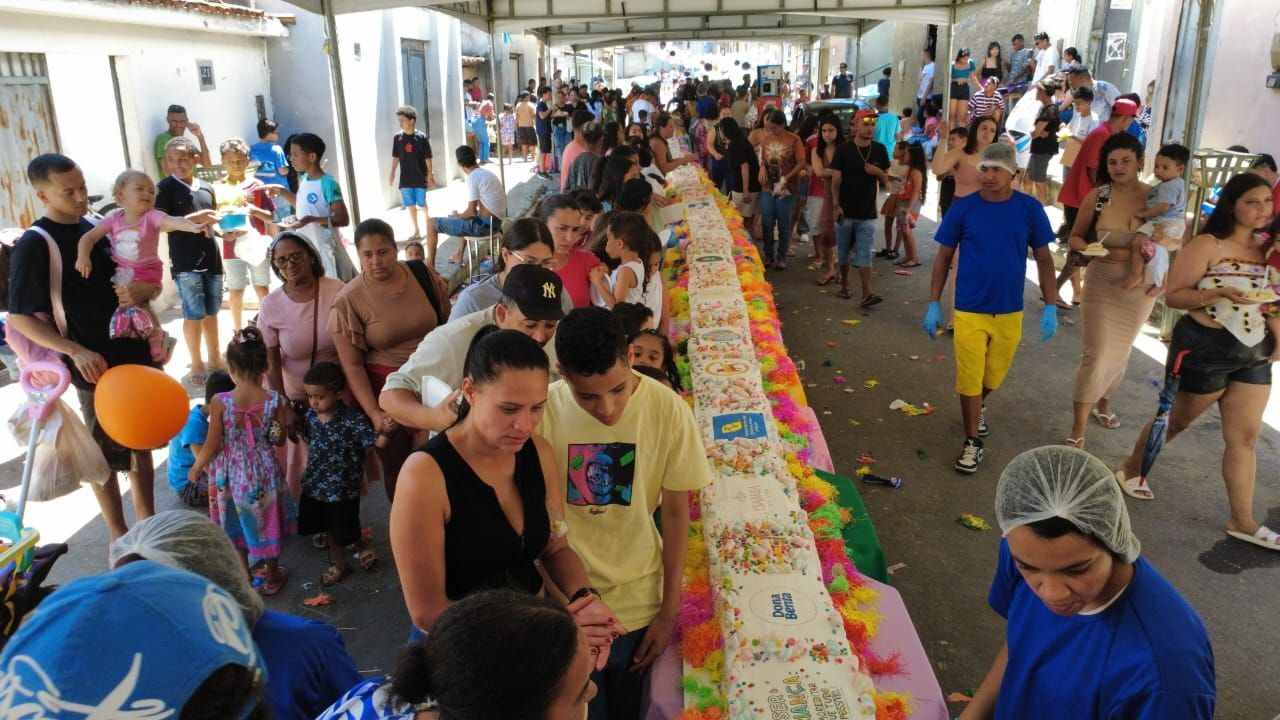 Crianças e adultos ao redor do bolo disposto sobre mesas durante o projeto Gigante Solidário no bairro Nossa Senhora Aparecida