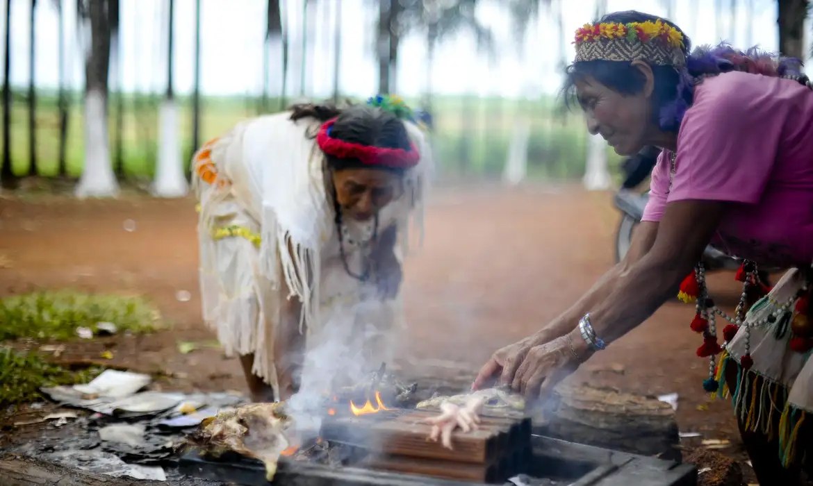 Duas pessoas com adornos de penas e colares junto a uma pequena fogueira ao ar livre. Foto de arquivo.