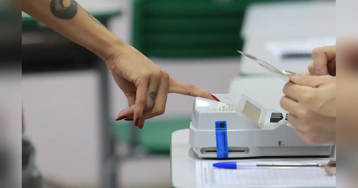 Detalhe de mãos junto a equipamento eletrônico e documentos em um local de votação