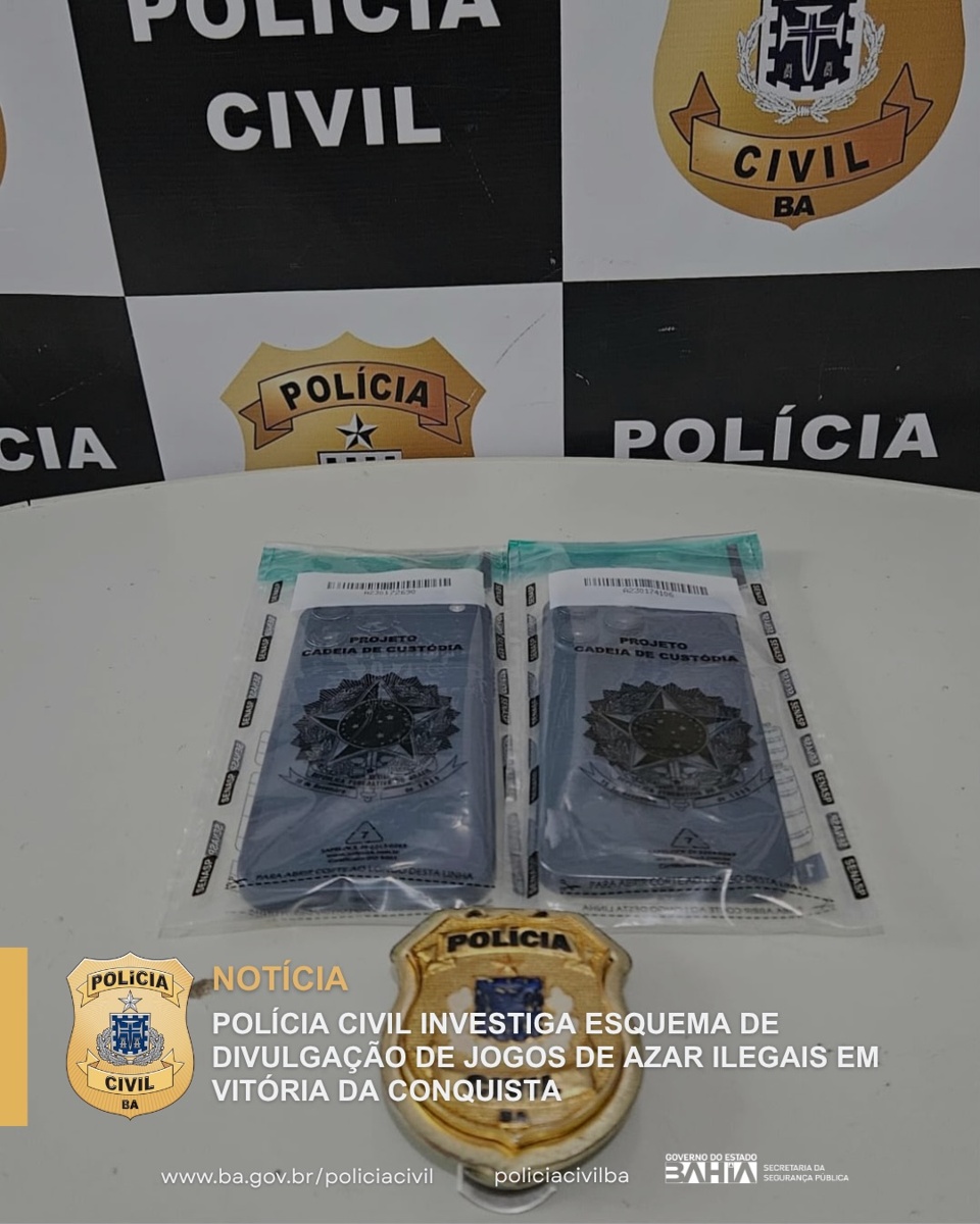 Dois celulares em embalagens de cadeia de custódia sobre mesa, diante de painel da Polícia Civil da Bahia