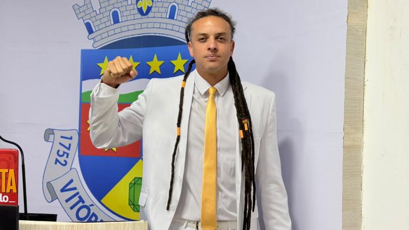 Homem de terno claro e gravata amarela, com o punho erguido diante do brasão municipal